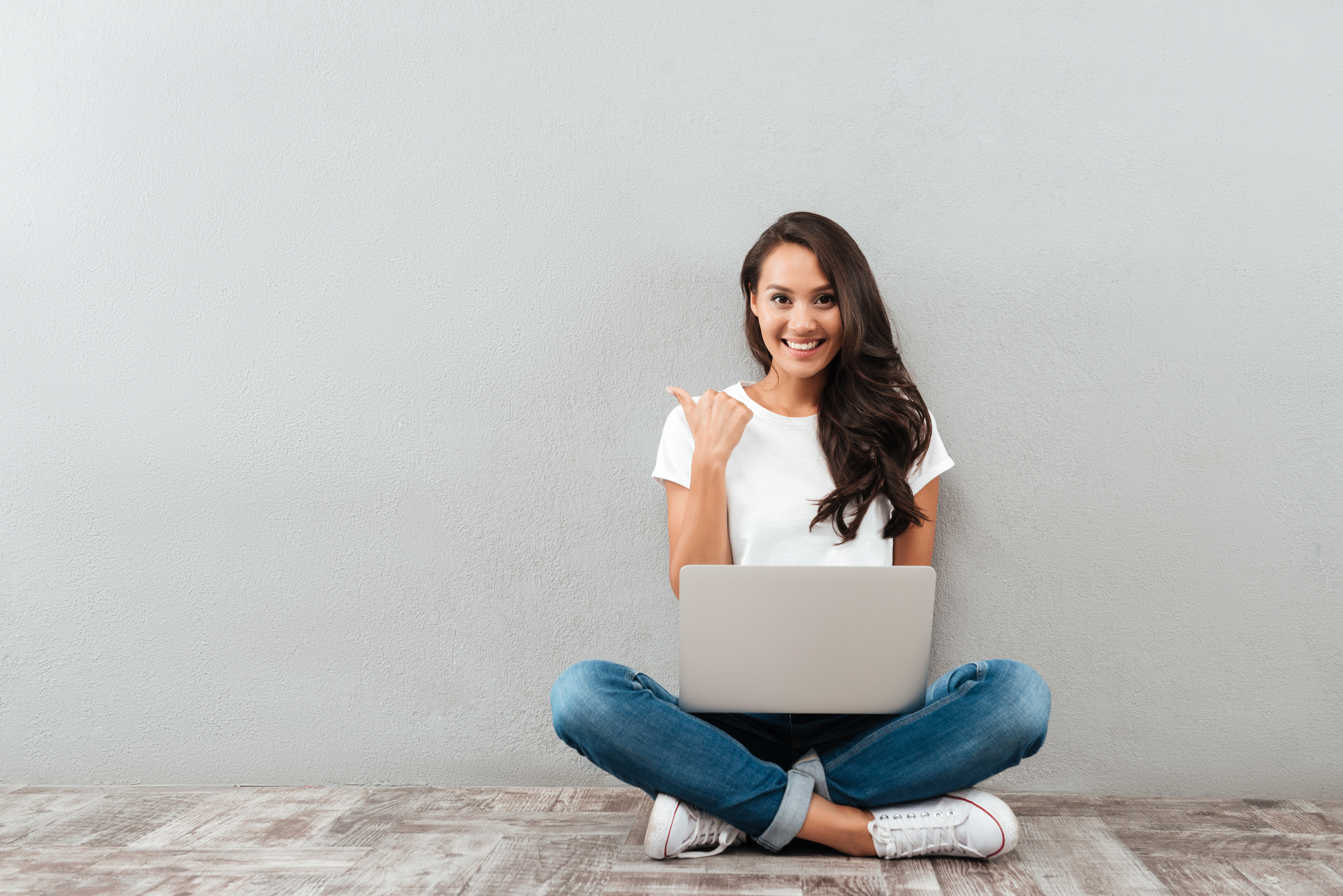 Happy smiling asian woman working on laptop computer while sitting on the floor with legs crossed and pointing finger away isolated over gray background